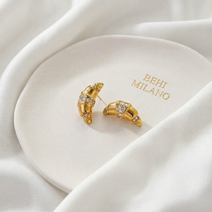 BEHI MILANO Aris Earrings — gold croissant studs displayed on white ceramic tray with BEHI MILANO logo
