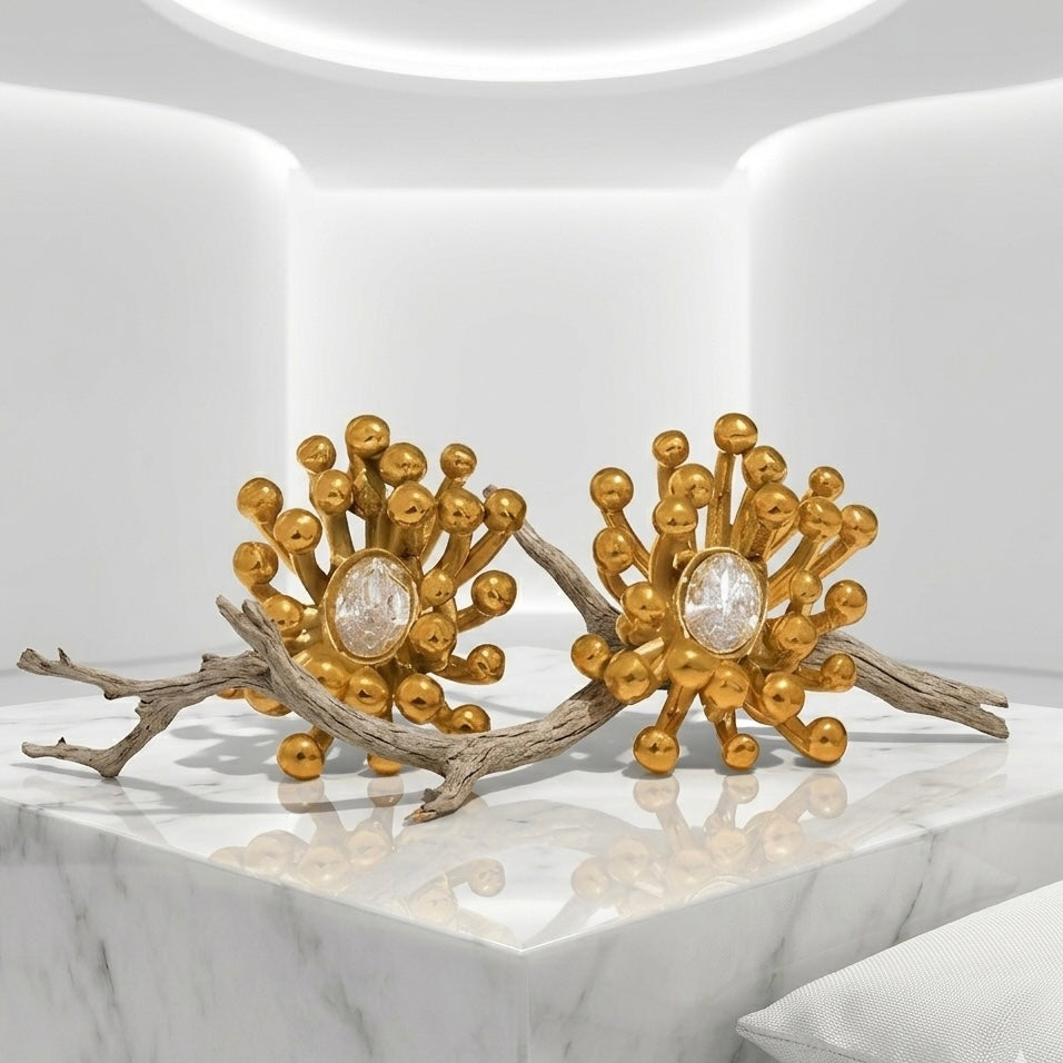 BEHI MILANO Artymanos Earrings — pair of 18k gold-plated crystal earrings placed on natural wood branch