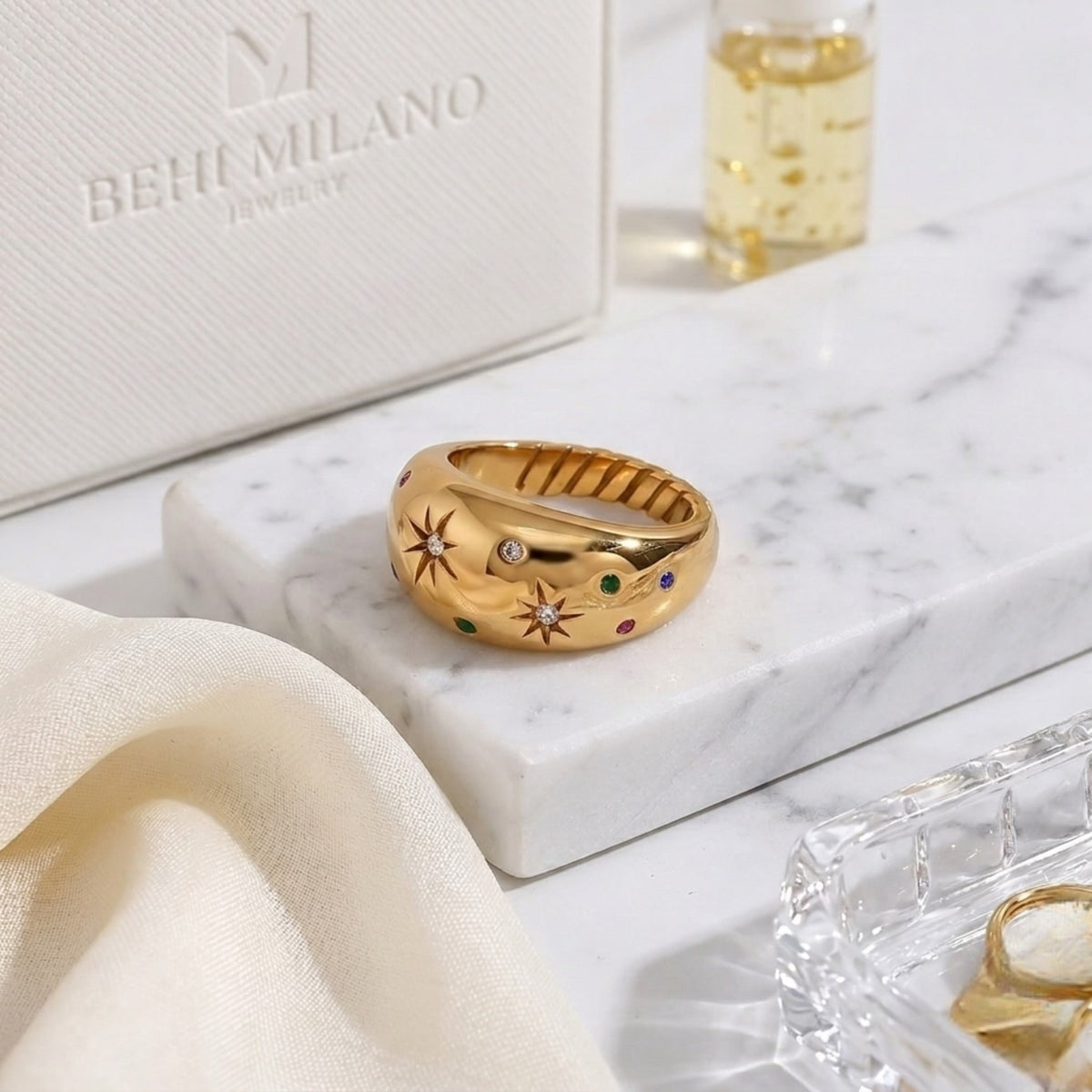 BEHI MILANO Camila Ring — gold colorful zircon ring displayed on white marble with luxury bottle
