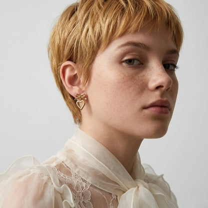 BEHI MILANO Cara Earrings — model with short blonde hair in sheer lace top wearing gold bow heart earrings