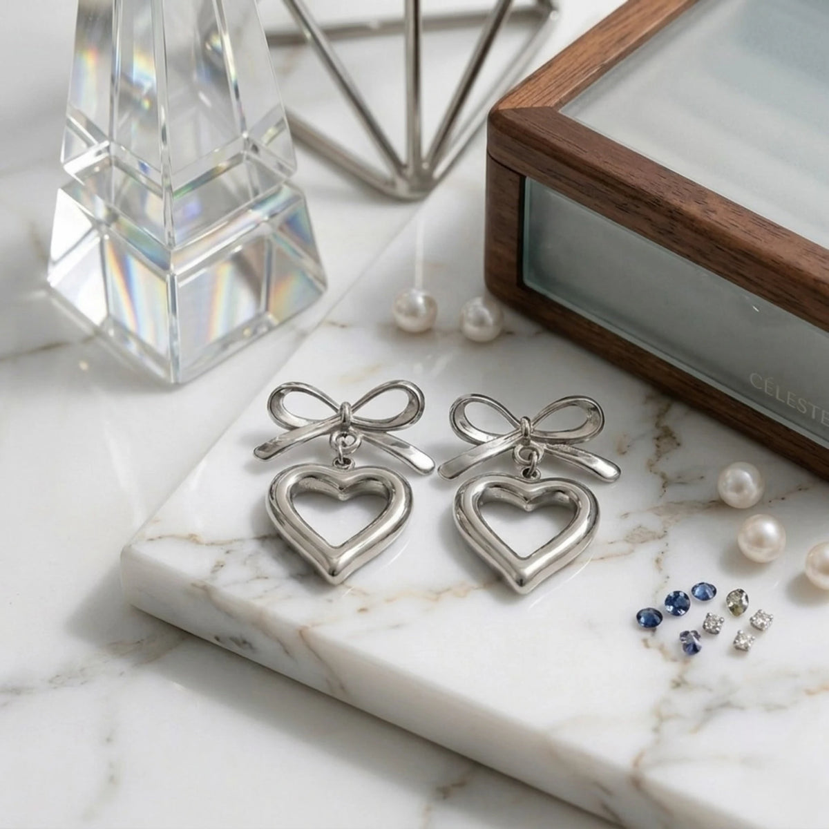 BEHI MILANO Cara Earrings — silver bow and open heart drop earrings on white marble