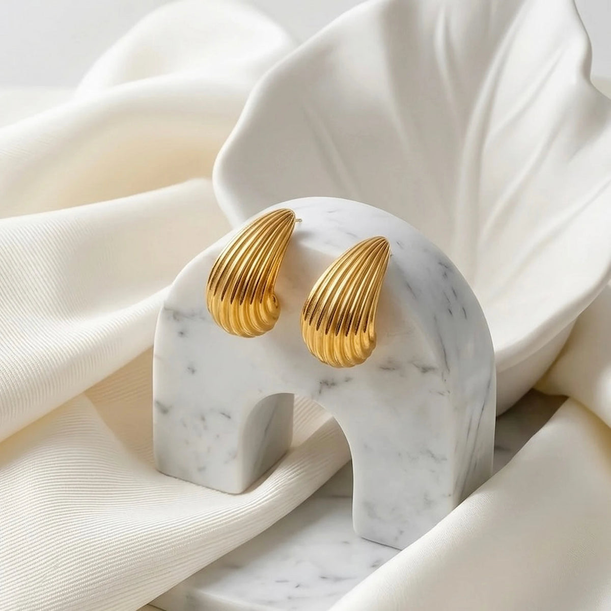 BEHI MILANO Claire Earrings — gold sculptural drop studs on white marble arch display