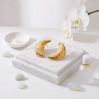 BEHI MILANO Croissant Earrings — gold crescent drop studs on square white marble pedestal with cosmetic bottle