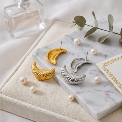 BEHI MILANO Croissant Earrings — gold and silver crescent studs on marble slab with freshwater pearls and eucalyptus