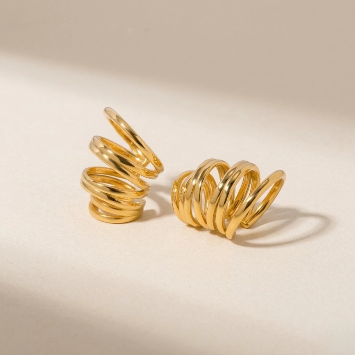 BEHI MILANO Denyse Ear Cuffs — 18k gold spiral ear cuffs resting on beige neutral background
