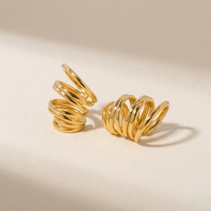 BEHI MILANO Denyse Ear Cuffs — 18k gold spiral ear cuffs resting on beige neutral background
