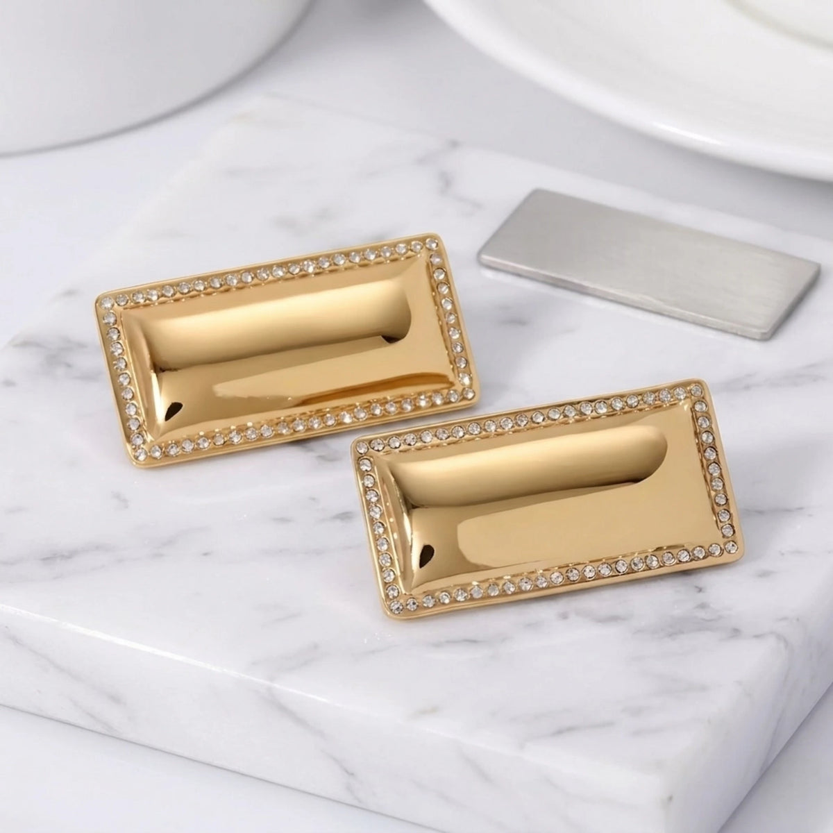 BEHI MILANO Dezi Earrings — pair of gold rectangular crystal-trimmed studs on white marble slab