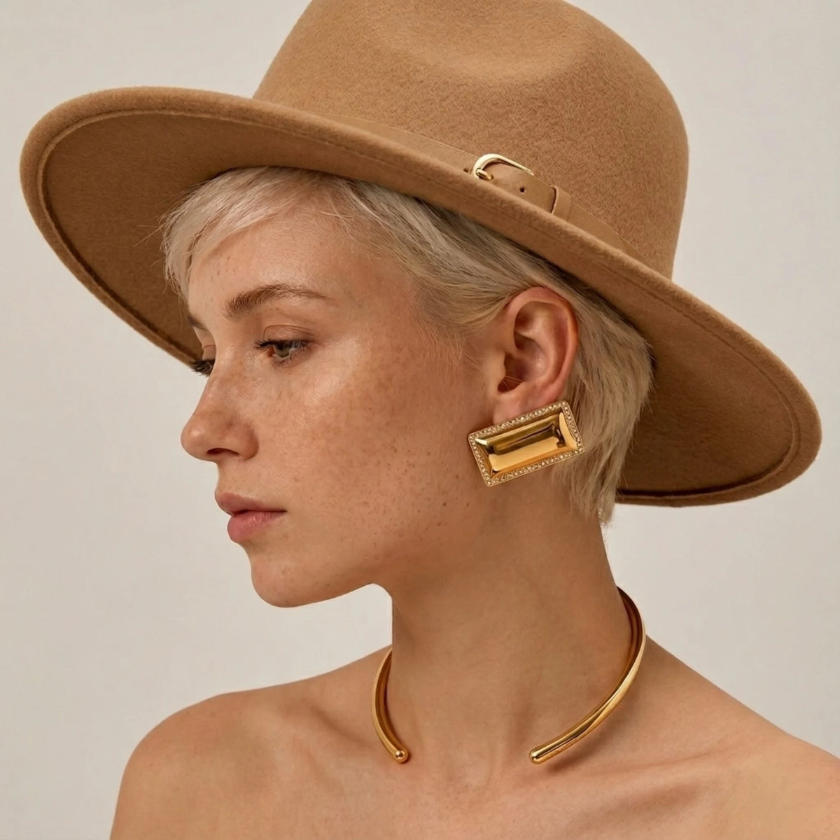 BEHI MILANO Dezi Earrings — model wearing oversized gold rectangular statement studs with tan fedora hat