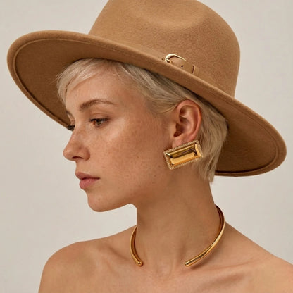 BEHI MILANO Dezi Earrings — model wearing oversized gold rectangular statement studs with tan fedora hat