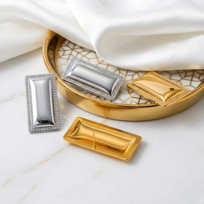 BEHI MILANO Dezi Earrings — gold and silver rectangular statement studs displayed on draped white silk