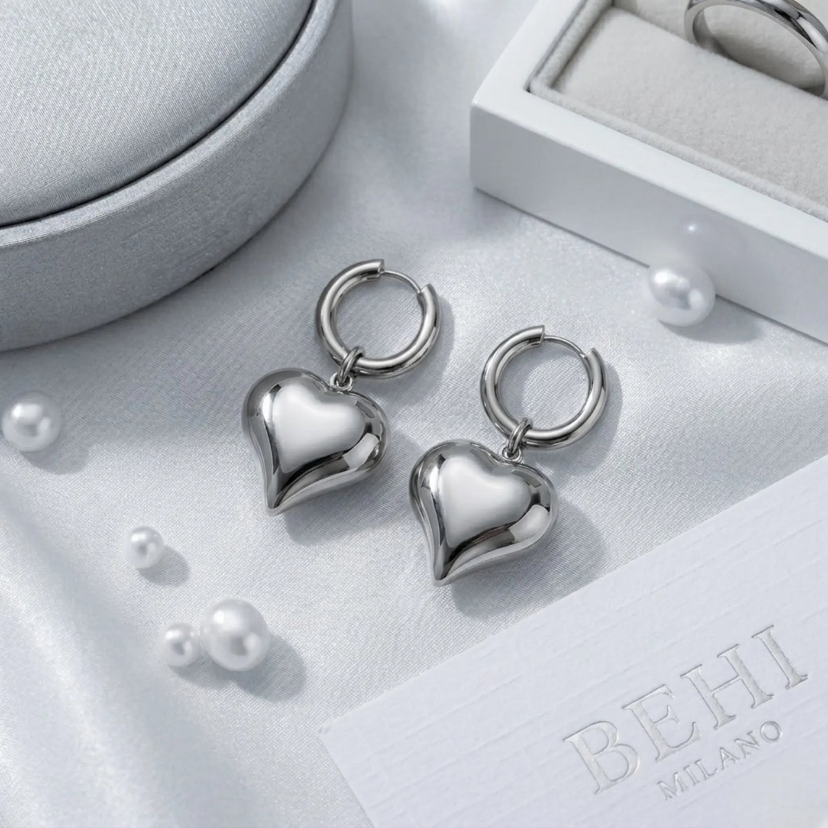 BEHI MILANO Emma Hoop Earrings — silver puffy heart drop hoop earrings on grey satin display tray