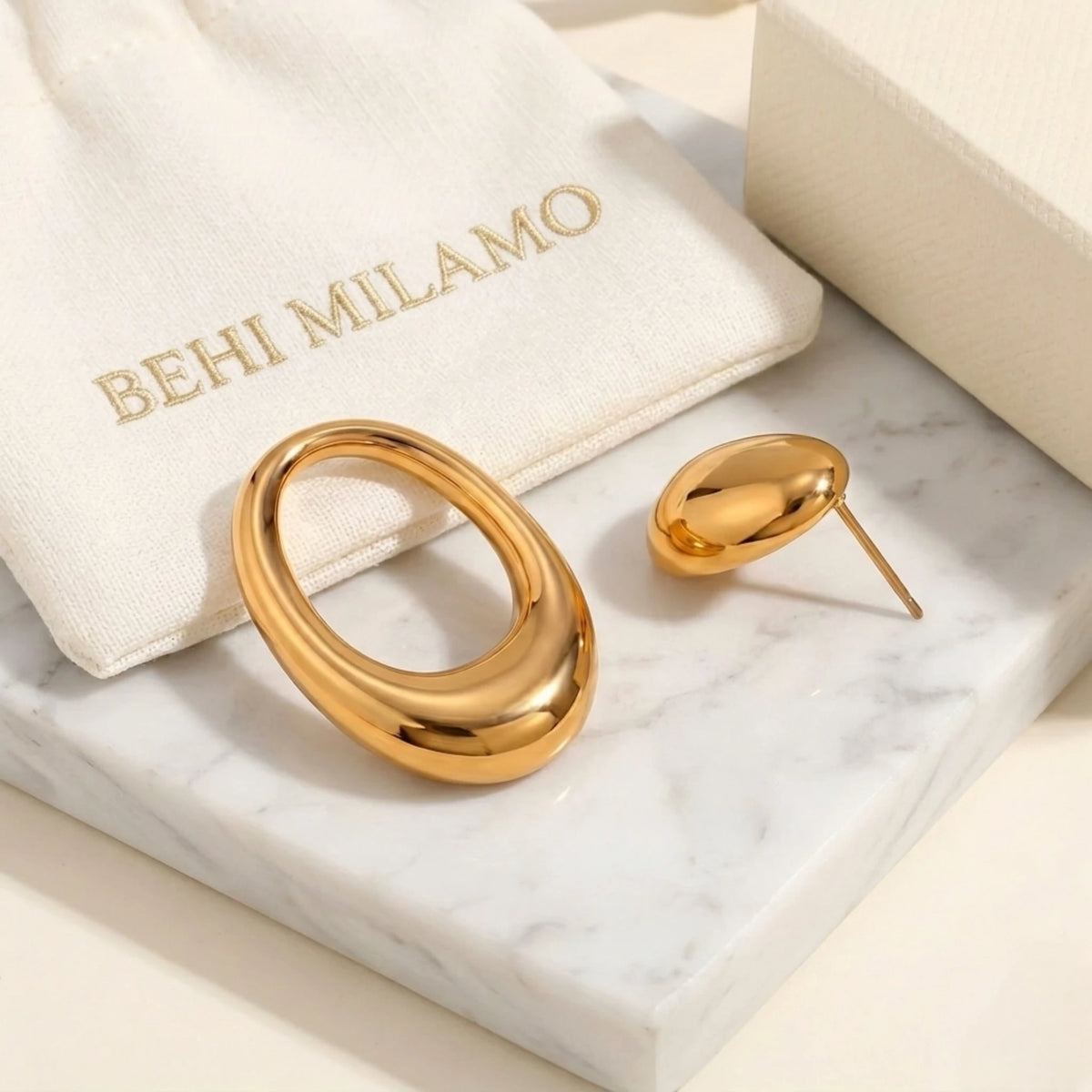BEHI MILANO Fiona Earrings — gold sculptural earrings next to signature BEHI MILANO pouch on marble