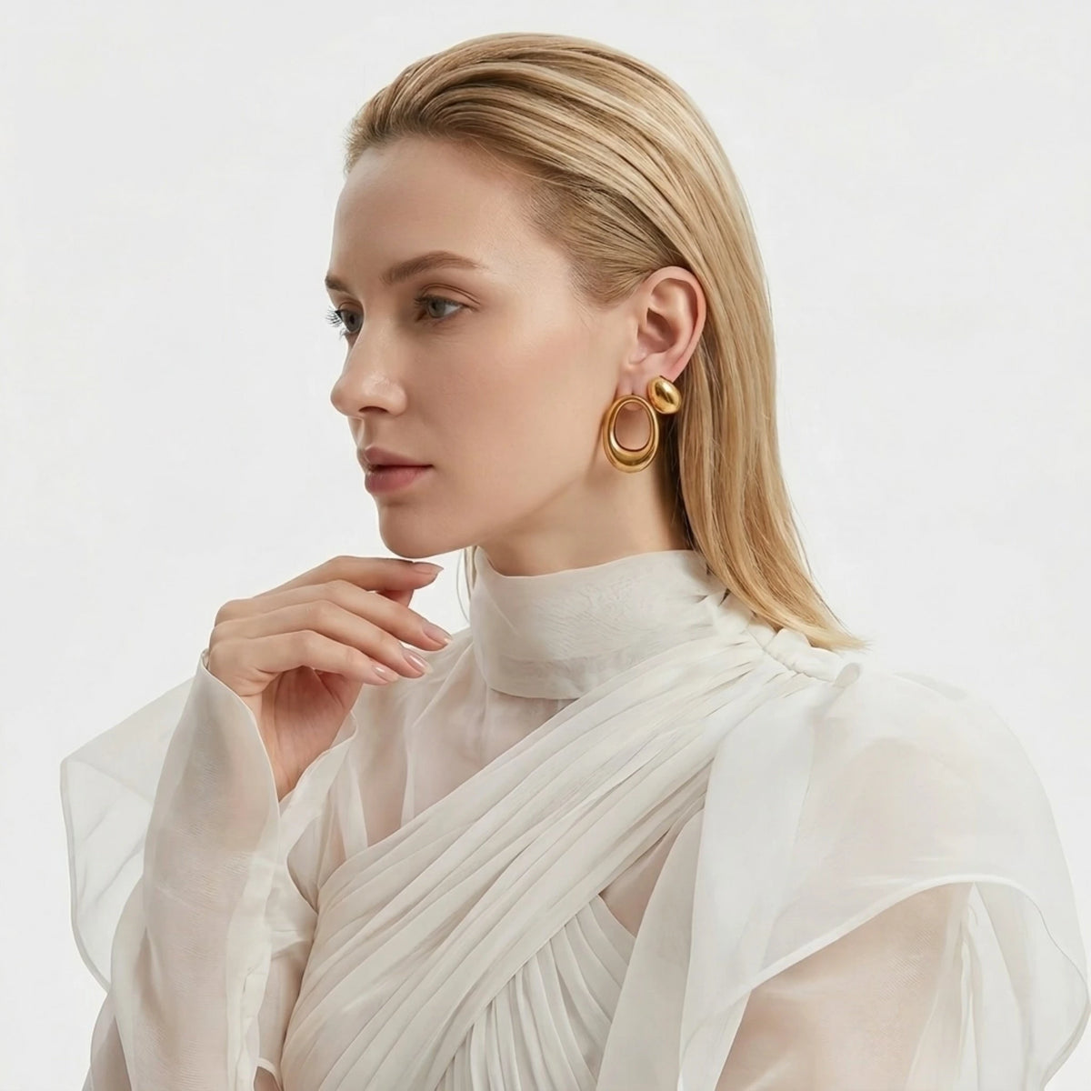 BEHI MILANO Fiona Earrings — blonde model wearing gold asymmetric sculptural earrings in sheer white top