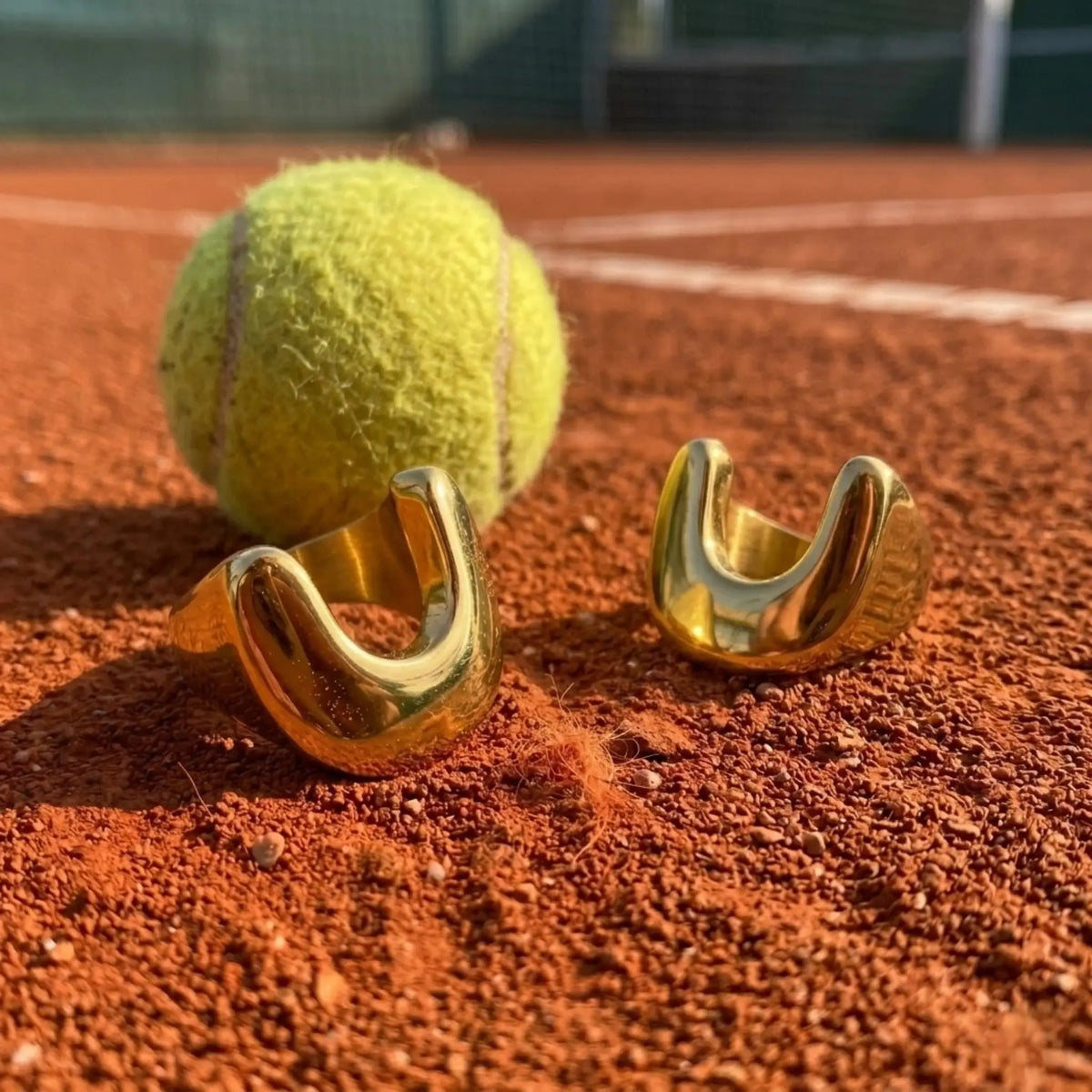 BEHI MILANO Giusy Ring — two 18k gold dipped rings displayed on red clay court near tennis ball