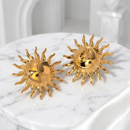BEHI MILANO Glow Earrings — gold solar motif statement studs resting on white marble pedestal