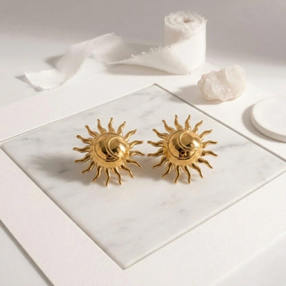 BEHI MILANO Glow Earrings — pair of gold sun studs displayed flat on white marble tray with silk ribbon