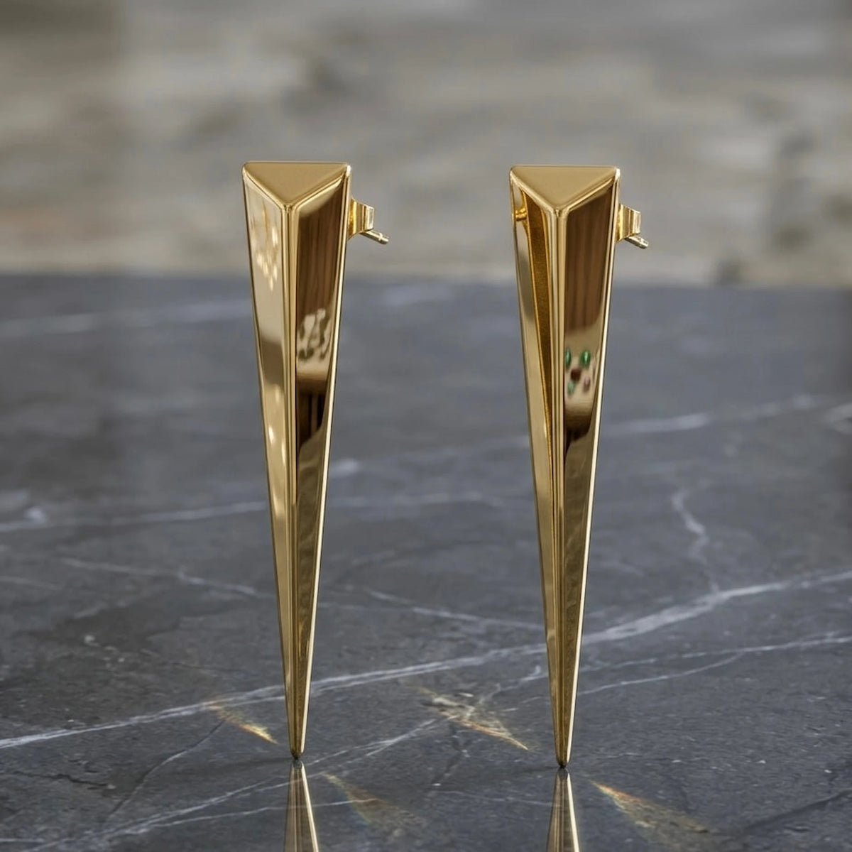BEHI MILANO Hanika Earrings — gold tapered geometric studs displayed on dark slate stone background