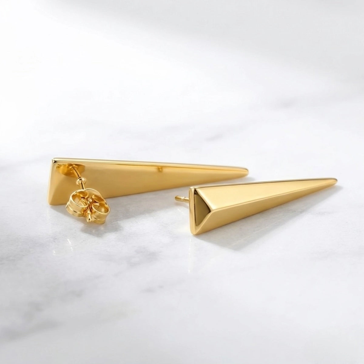 BEHI MILANO Hanika Earrings — pair of gold geometric drop studs resting flat on white marble surface