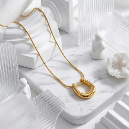 BEHI MILANO Horseshoe Necklace — 18k gold pendant necklace resting on white marble with sheer ribbon