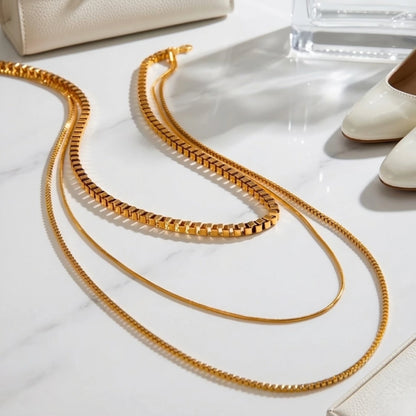 BEHI MILANO Ira Necklace — triple layered gold necklace laid flat on white marble next to high heels