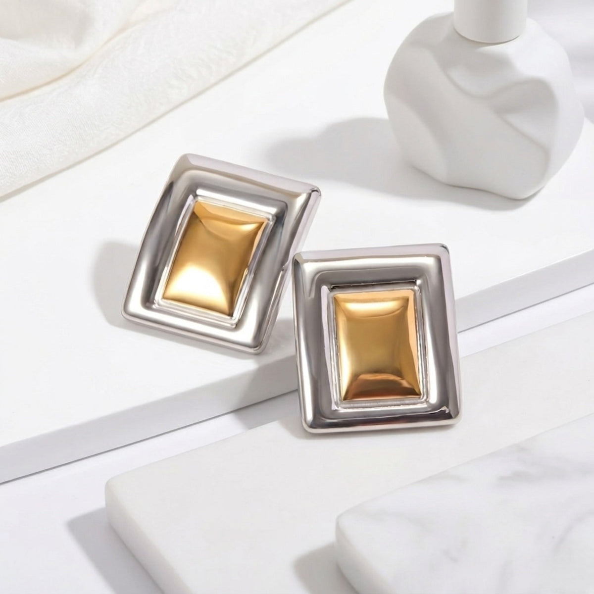 BEHI MILANO Isabella Earrings — two tone gold and silver studs resting on white geometric display podium
