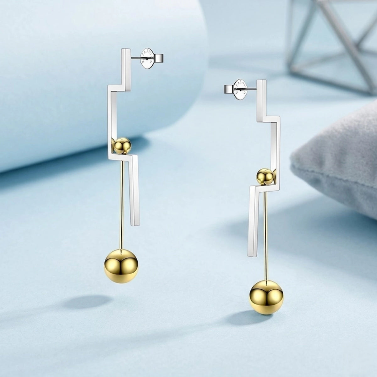 BEHI MILANO Isidora Earrings — two tone drop earrings on pastel blue geometric display background