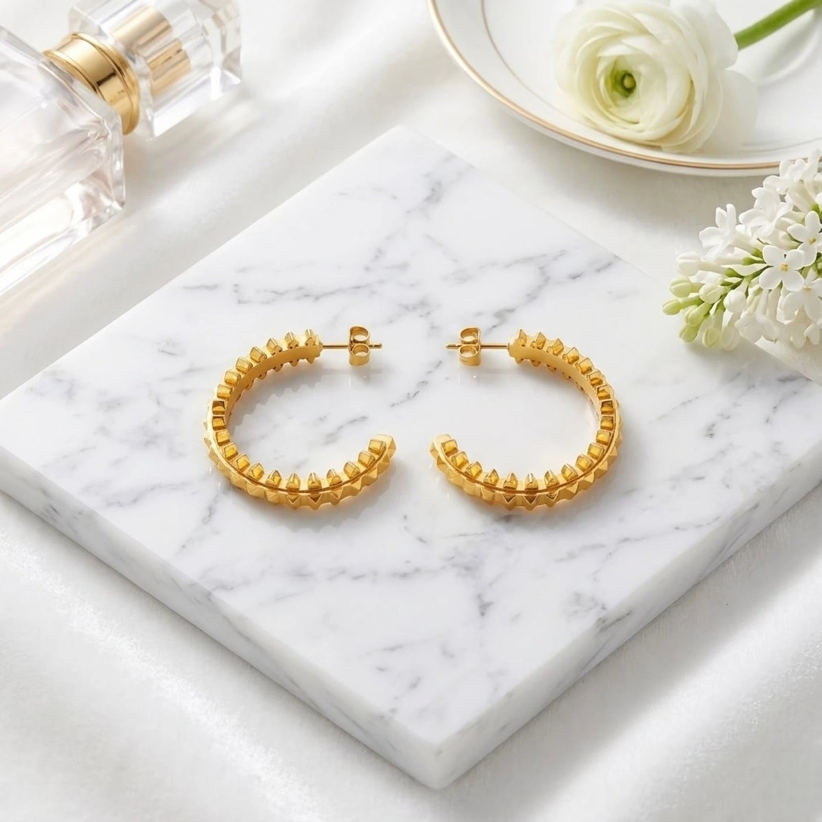 BEHI MILANO Isla Earrings — pair of 18k gold pyramid studded hoops on marble slab with white flowers
