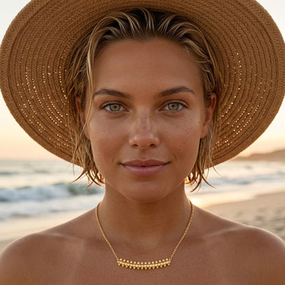 BEHI MILANO Isla Necklace — model wearing gold bar pendant necklace and straw sun hat on beach