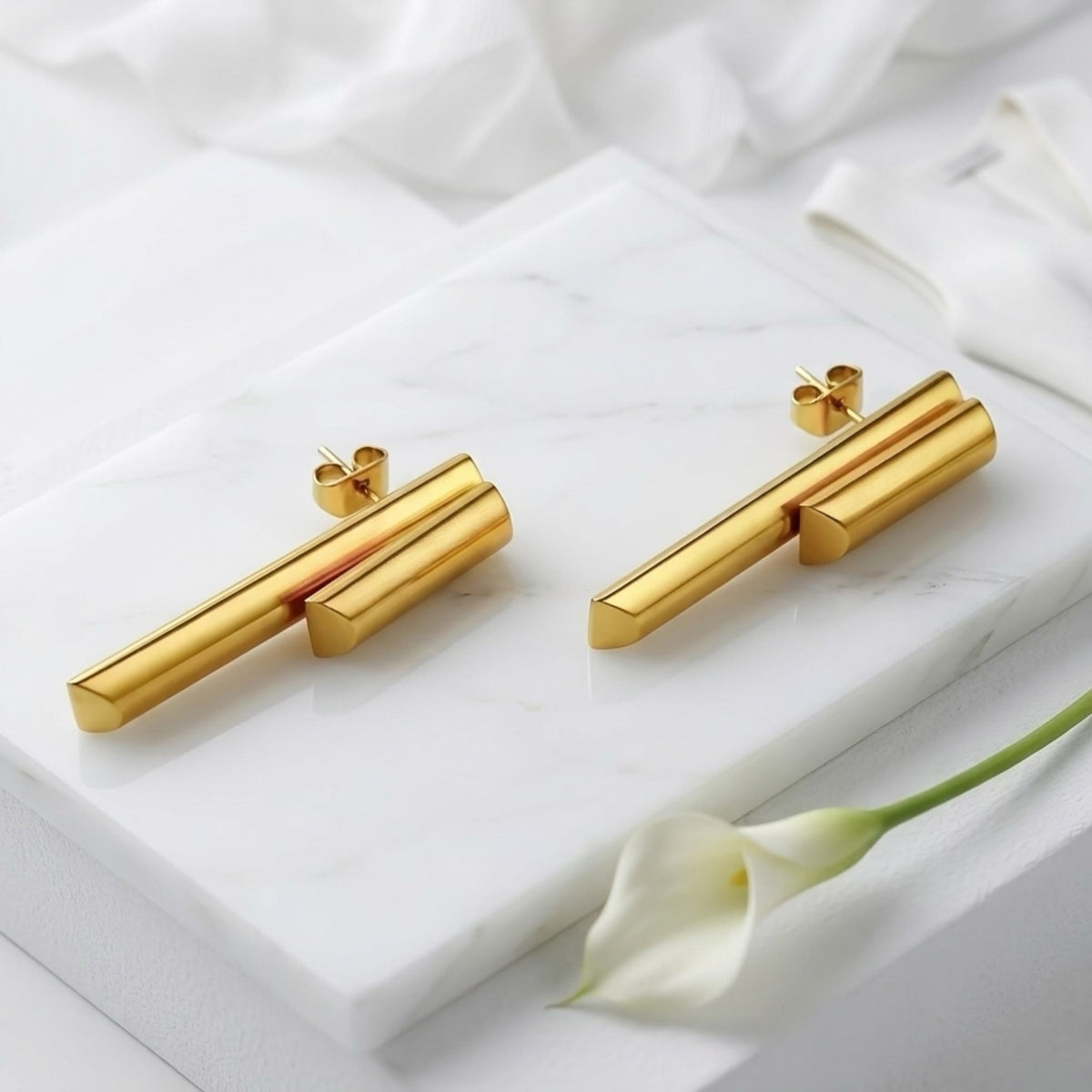 BEHI MILANO Ivana Earrings — gold geometric drop earrings displayed on white marble slab with floral accent