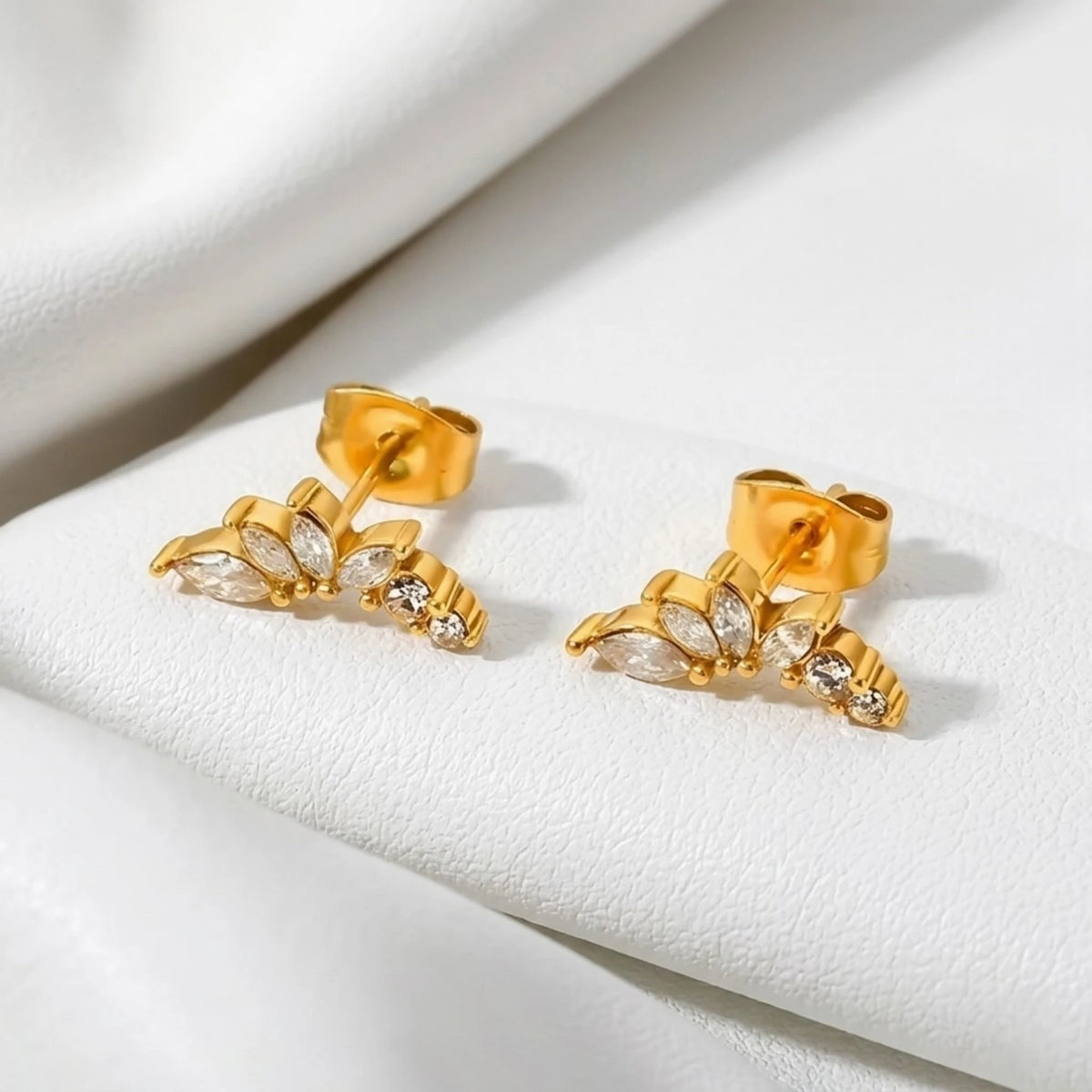 BEHI MILANO Josa Earrings — 18k gold crystal stud earrings resting on white satin fabric