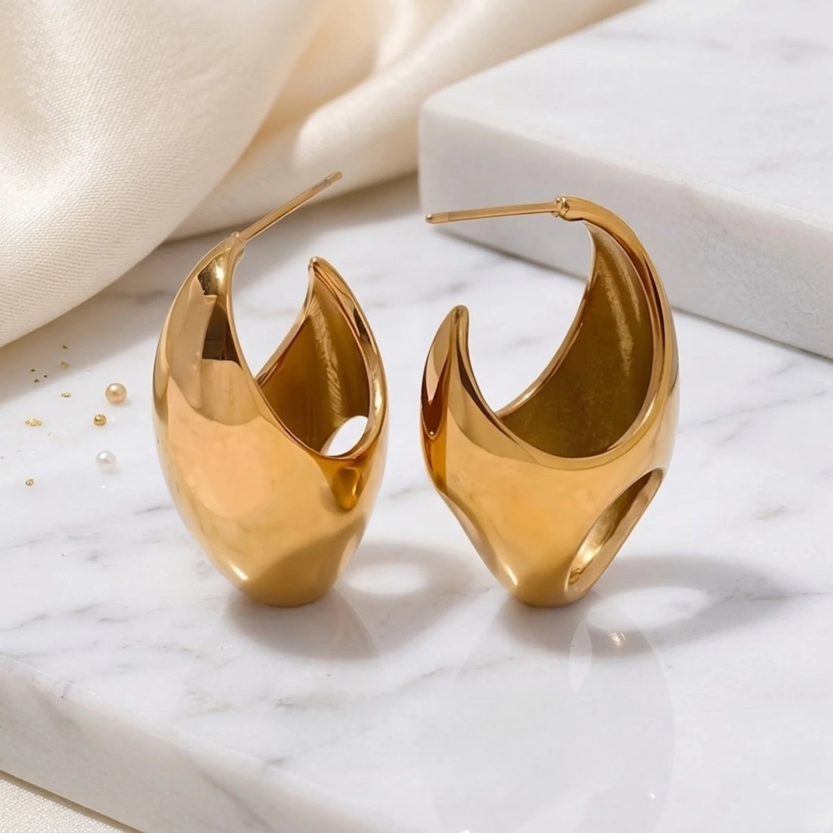 BEHI MILANO Judy Earrings — pair of gold teardrop open hoop earrings resting on marble surface