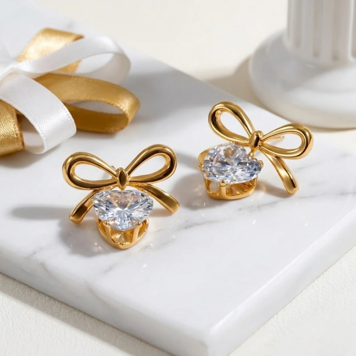 BEHI MILANO Juliette Earrings — pair of gold bow heart drop studs on white marble platform