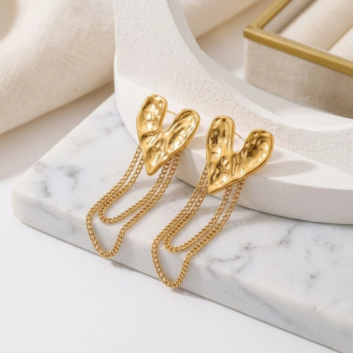 BEHI MILANO Ledda Earrings — gold hammered heart chain drop studs on white marble pedestal