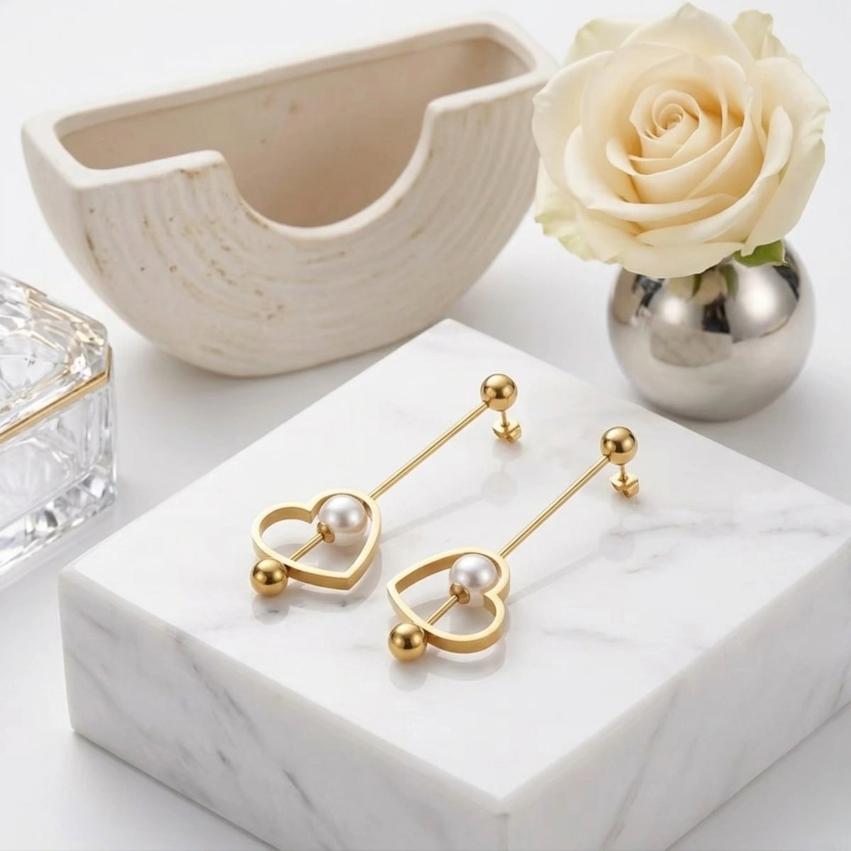BEHI MILANO Leona Earrings — gold heart pearl drop earrings on white pedestal with ceramic bowl and white rose